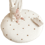 Baby sitting on a round cream-colored padded mat with small brown bear faces, dressed in a cozy bear-themed outfit including a white hat with bear ears, beige vest, and matching white clothing. A coordinating blanket is draped over a beige sofa behind the baby, with a knitted bear-face pillow placed nearby. Isolated on a transparent background for clean, stylish presentation